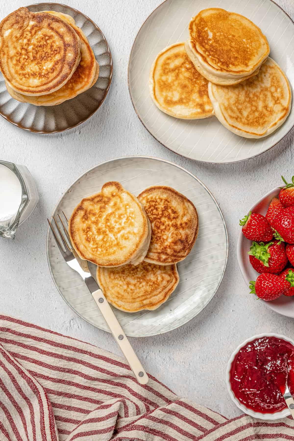 Overhead view of plates of gluten-free pikelets next to a bowl of strawberries and various toppings.