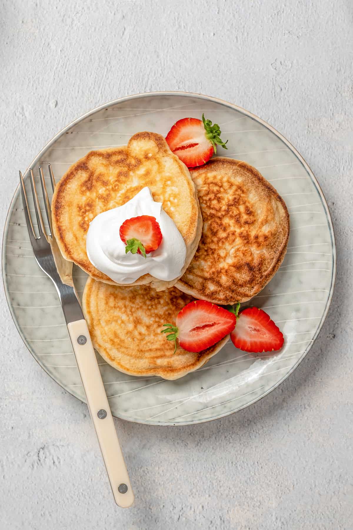 Overhead view of three gluten-free pikelets on a plate topped with whipped cream and strawberry slices.