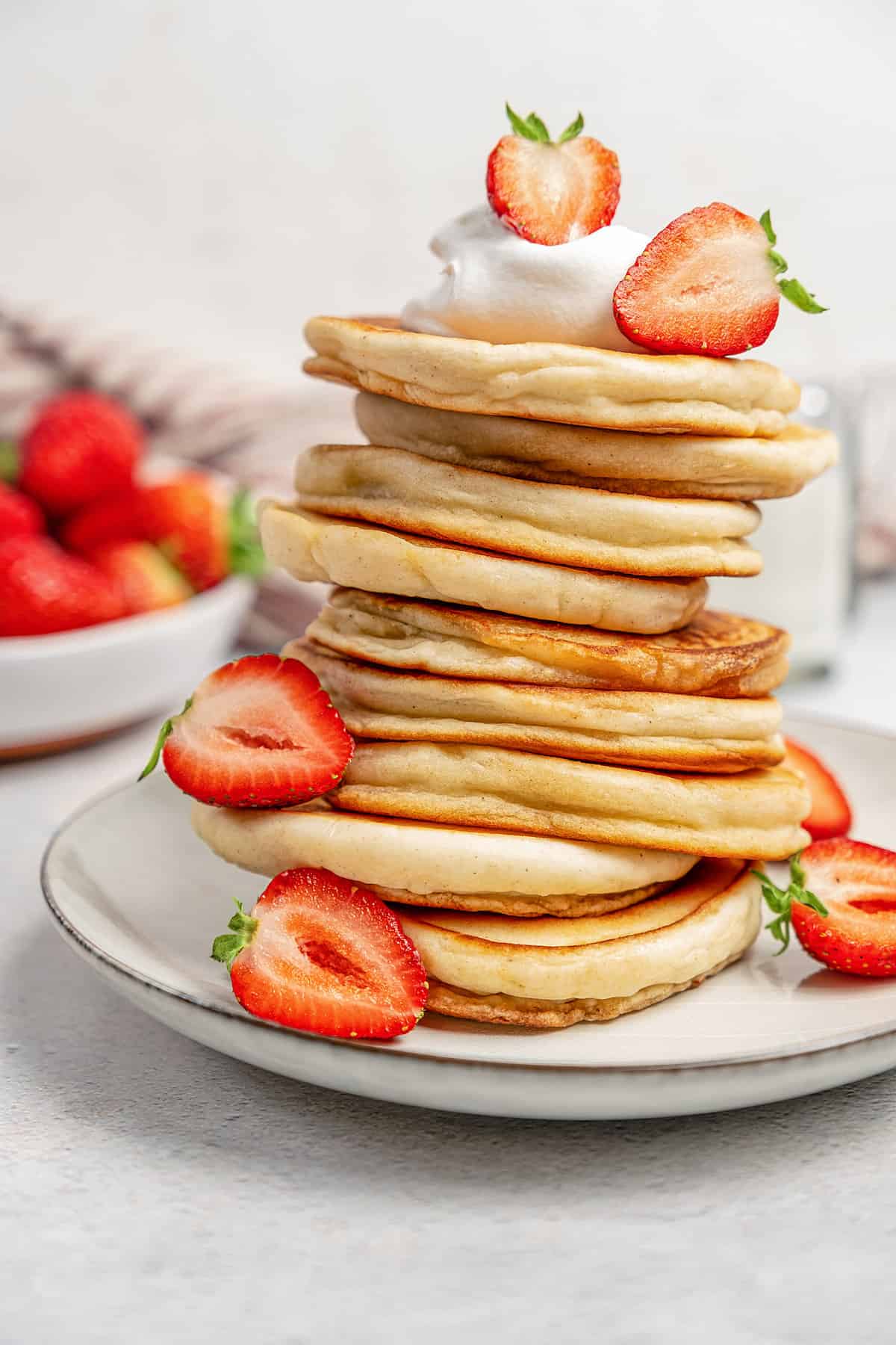 A tall stack of gluten-free pikelets on a plate garnished with whipped cream and sliced strawberries.