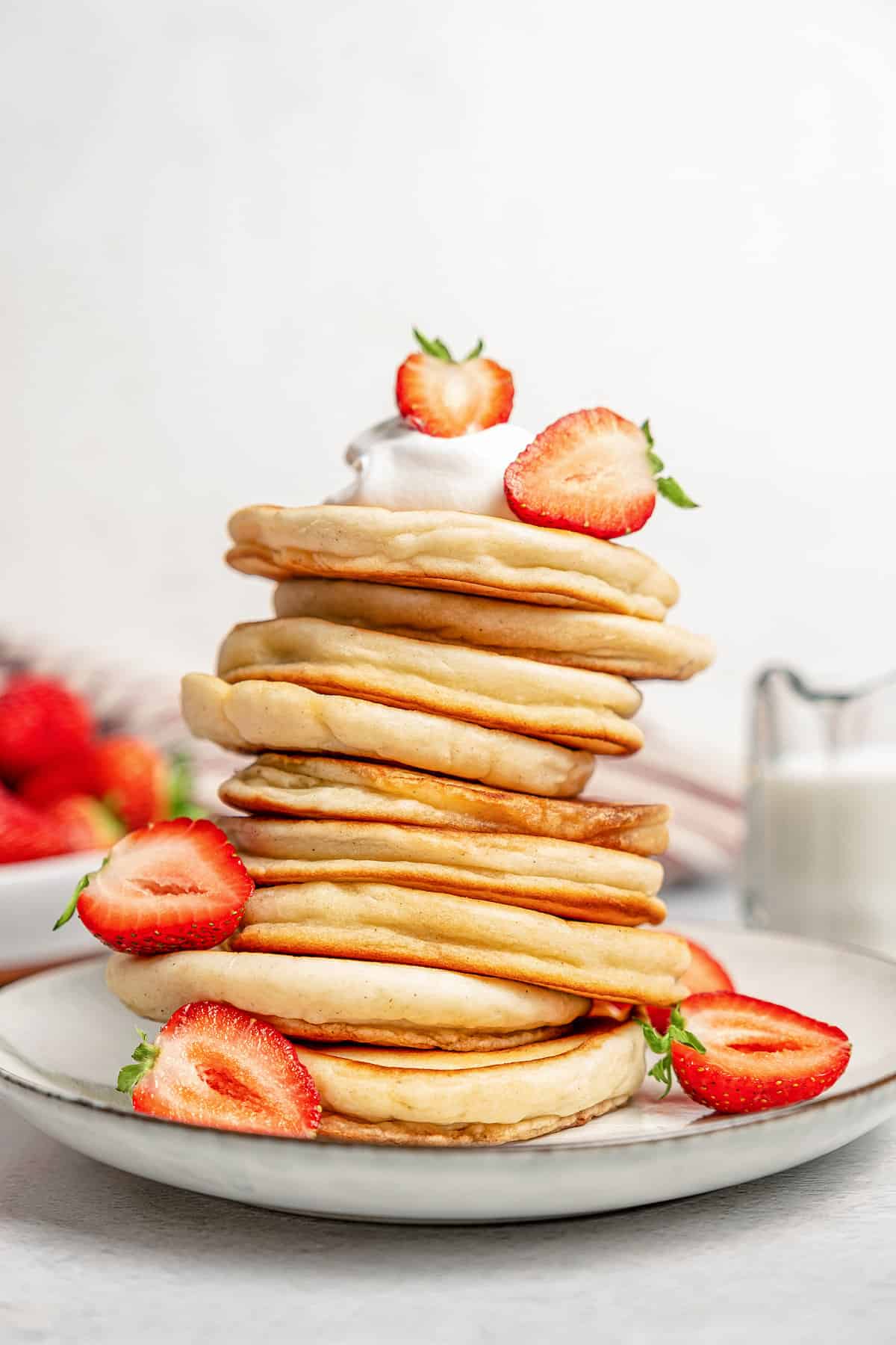 A tall stack of gluten-free pikelets on a plate garnished with whipped cream and sliced strawberries.