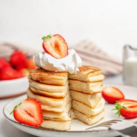 A stack of gluten-free pikelets on a plate garnished with whipped cream and sliced strawberries, with a slice missing.