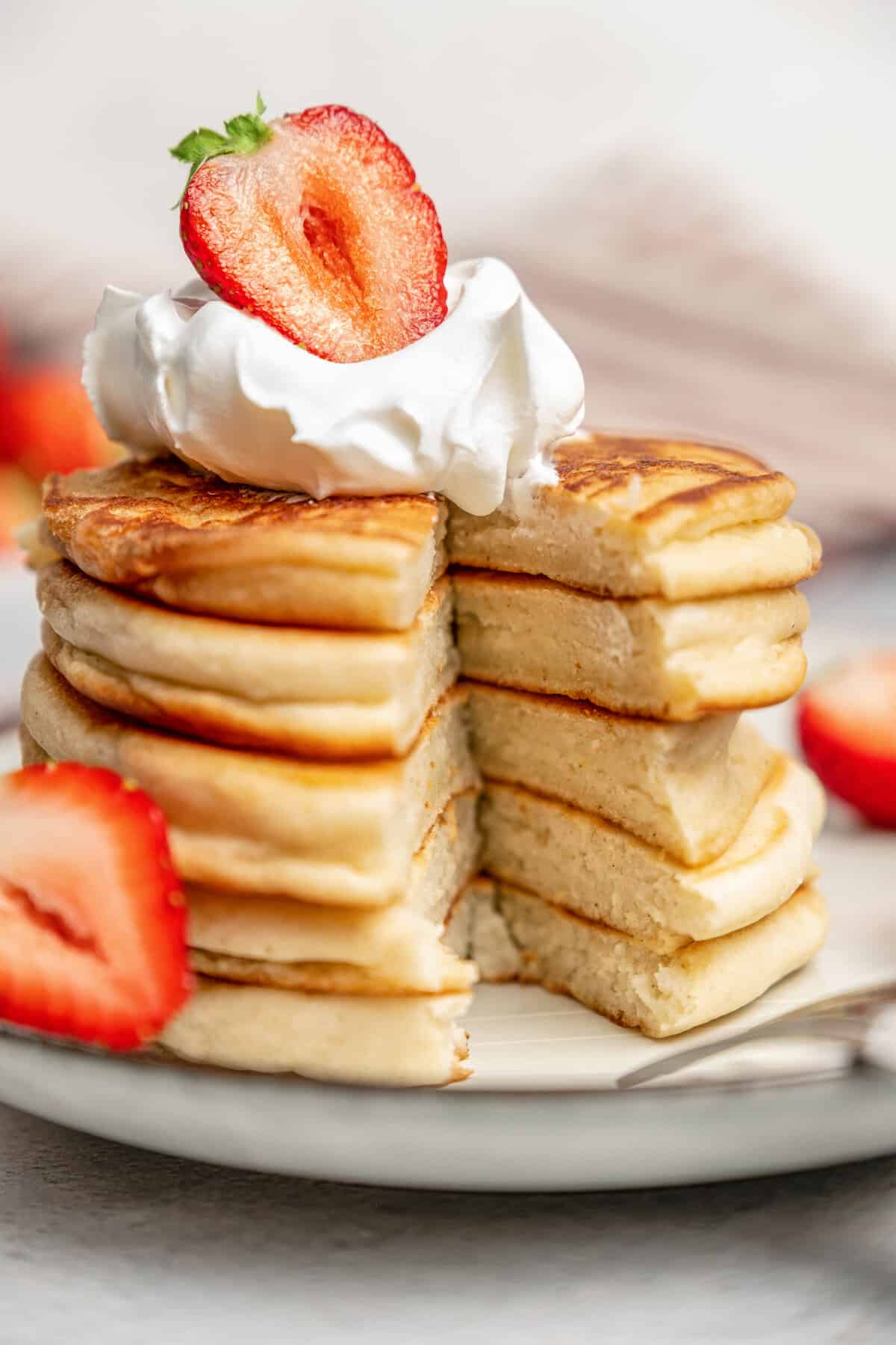 A stack of gluten-free pikelets on a plate garnished with whipped cream and sliced strawberries, with a slice missing.