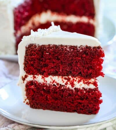 A slice of gluten-free red velvet cake with cream cheese frosting on a white plate with the rest of the cake in the background.
