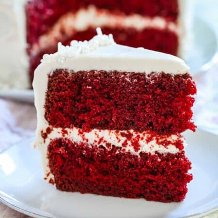 A slice of gluten-free red velvet cake with cream cheese frosting on a white plate with the rest of the cake in the background.