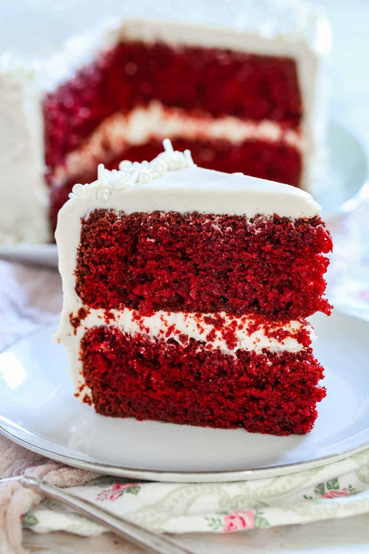 A slice of gluten-free red velvet cake with cream cheese frosting on a white plate with the rest of the cake in the background.