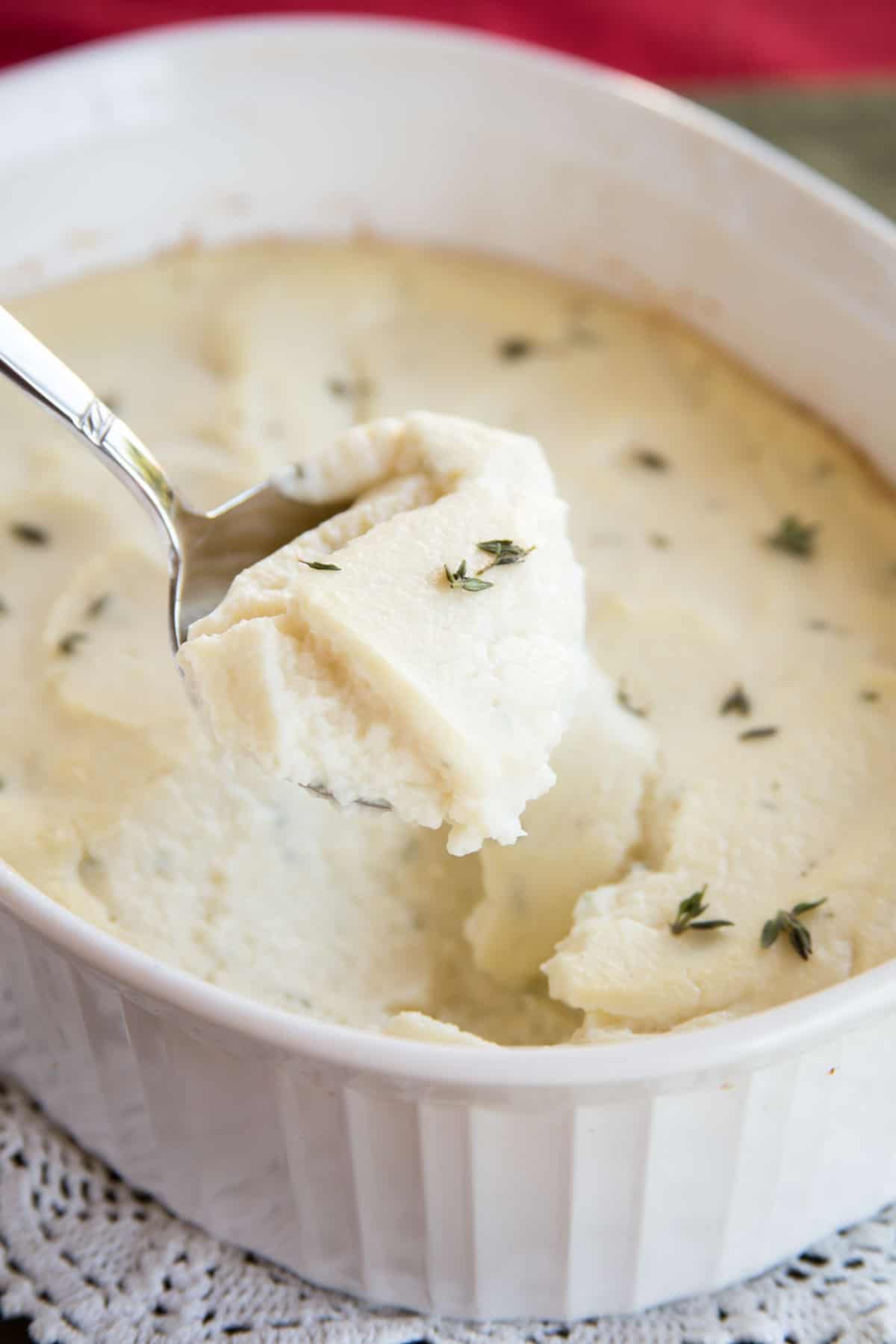 Goat cheese whipped cauliflower in a white casserole dish garnished with thyme being scooped up with a serving spoon.