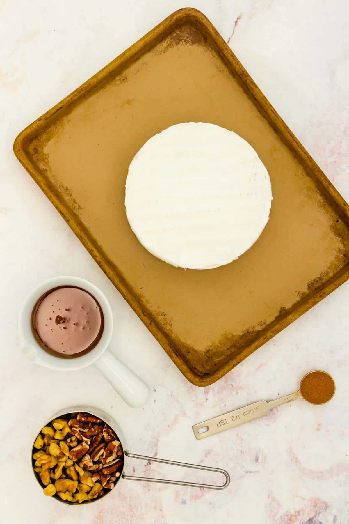 A wheel of Brie cheese, a measuring cup of walnuts and pecans, a half teaspoon of cinnamon, and a small dish of honey on a marble countertop.