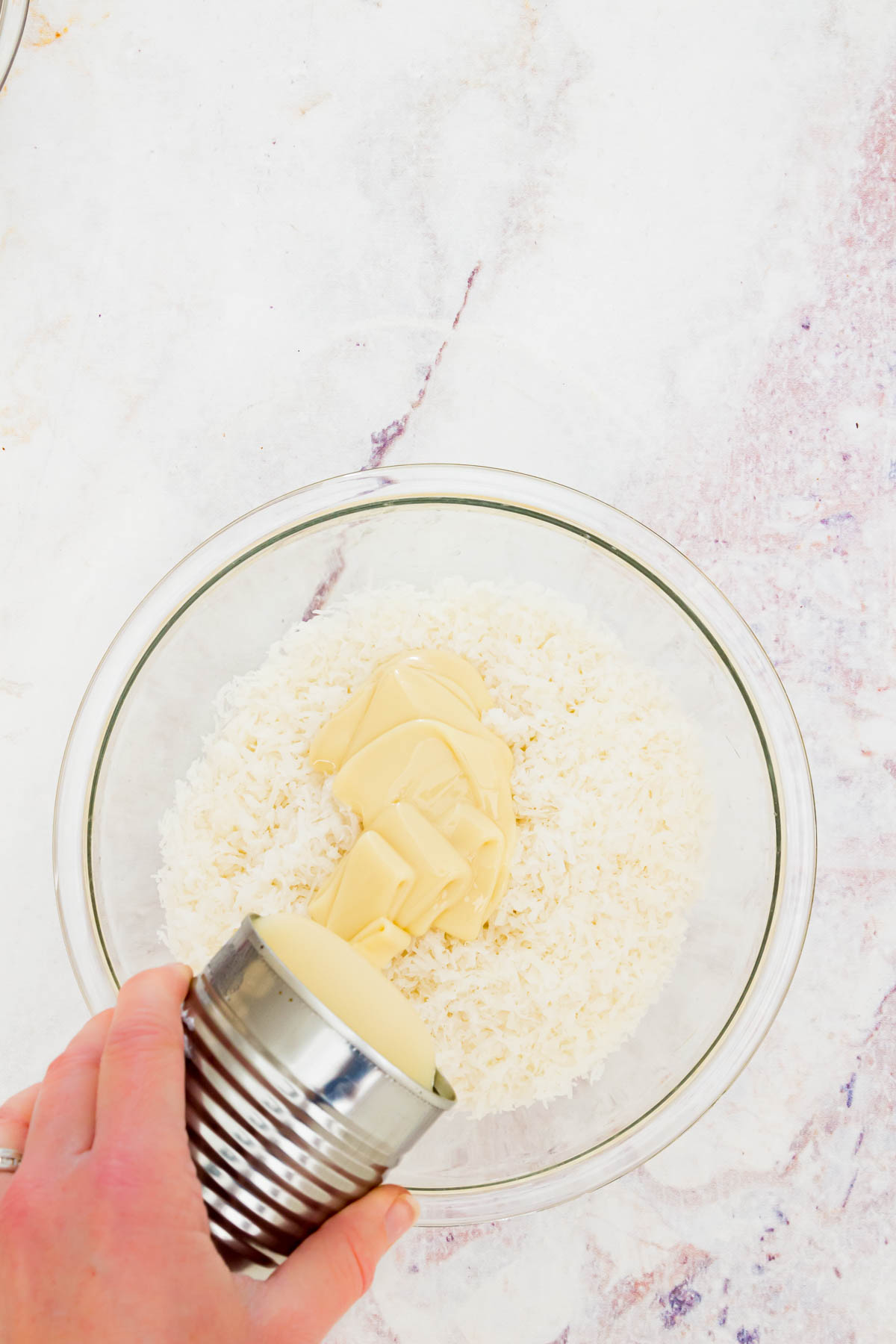 A can of sweetened condensed milk being poured over shredded coconut in a glass bowl.