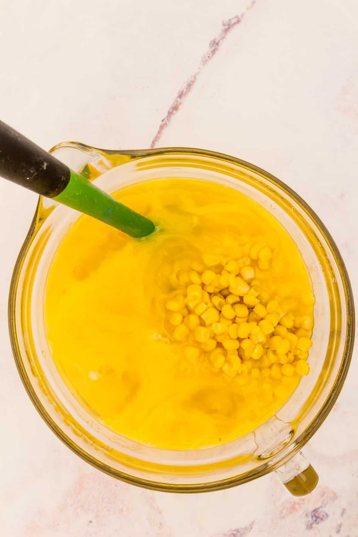 A green spatula in a glass mixing bowl with corn kernels and beaten eggs.