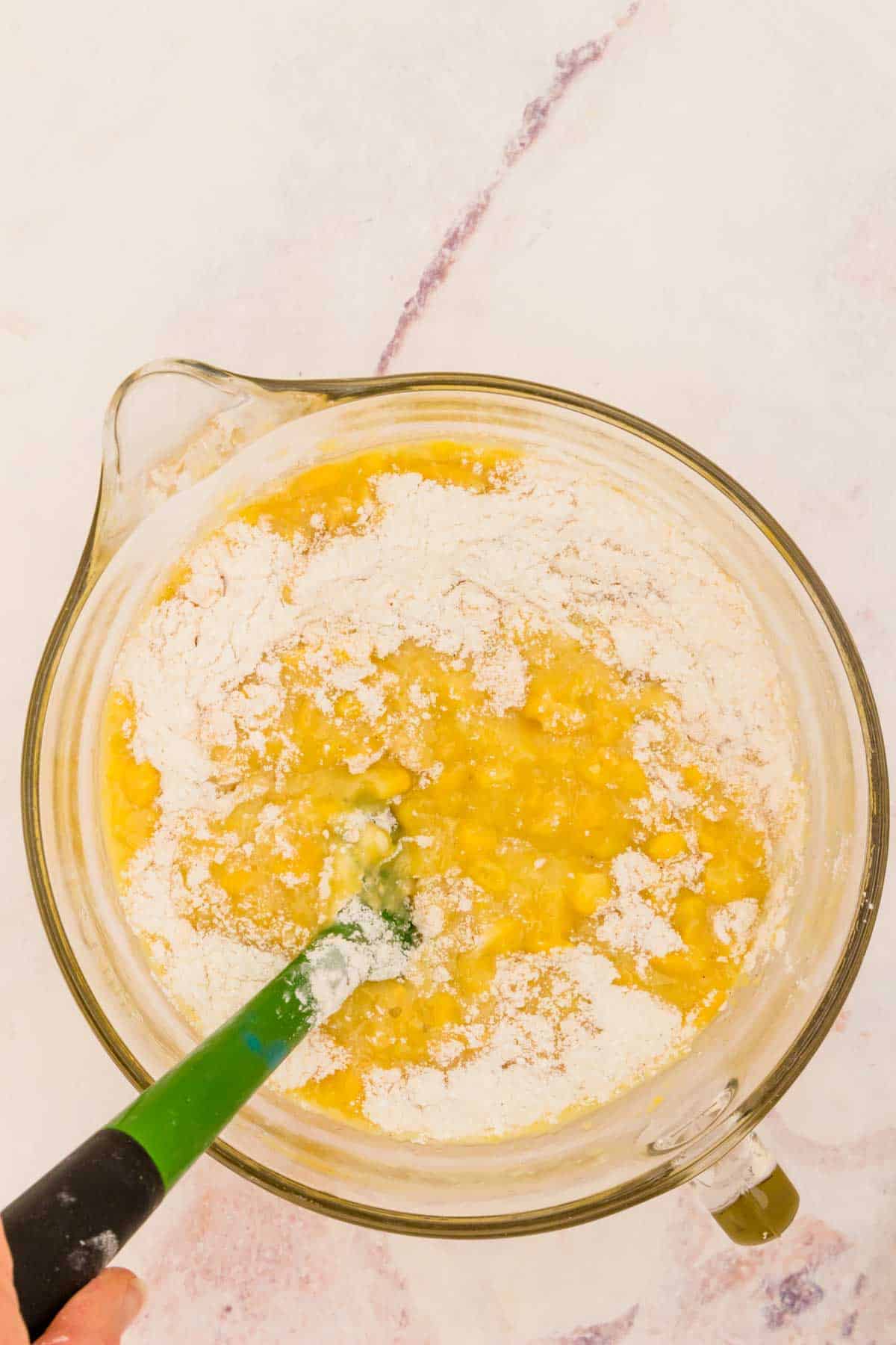 Dry ingredients being folded into the corn and egg mixture in a glass bowl with a green spatula.