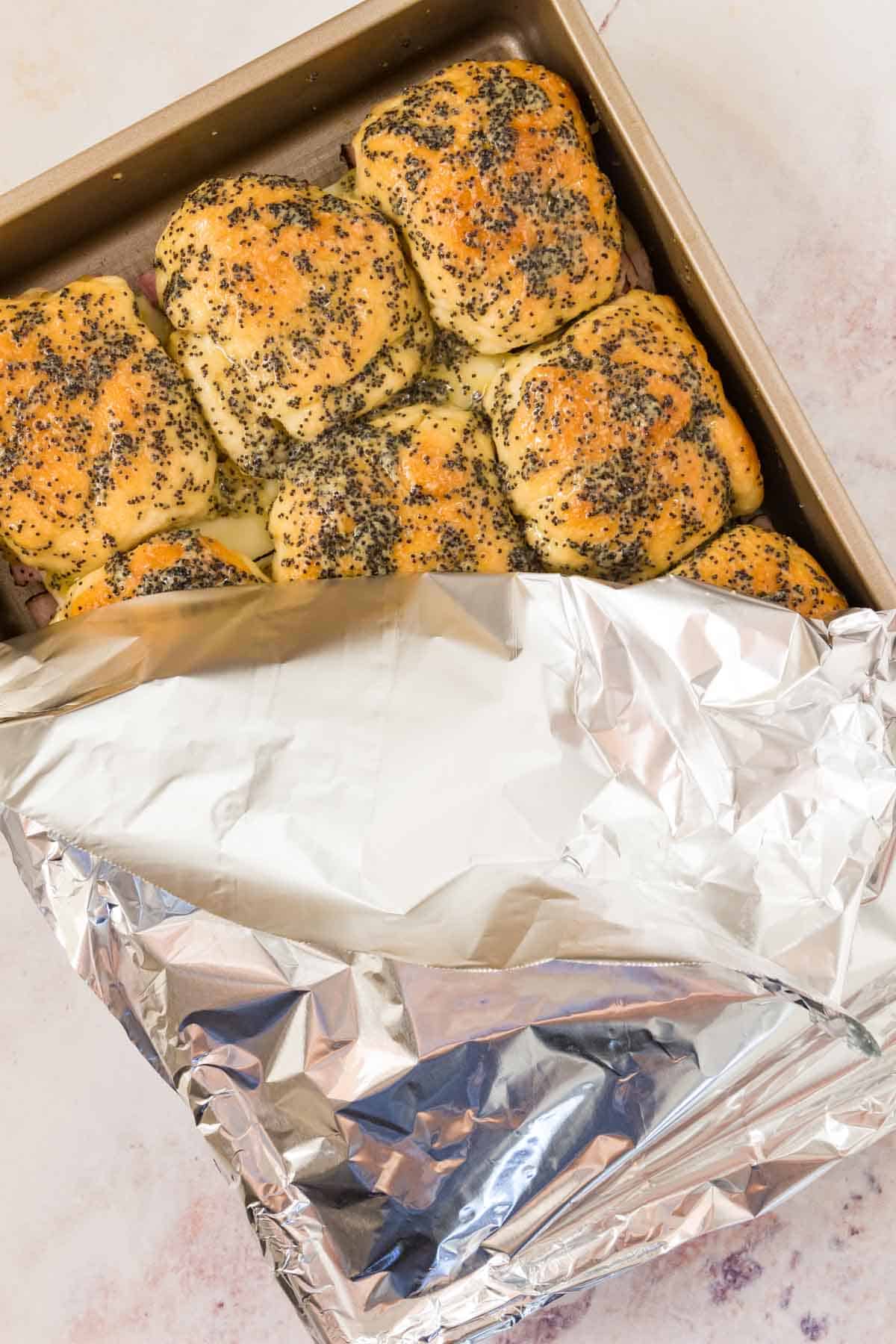 A pan of gluten-free ham and cheese sliders partially covered with aluminum foil.
