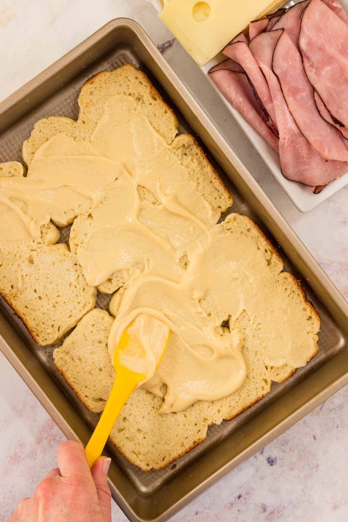 Mustard is spread over a layer of Hawaiian rolls in a baking pan.