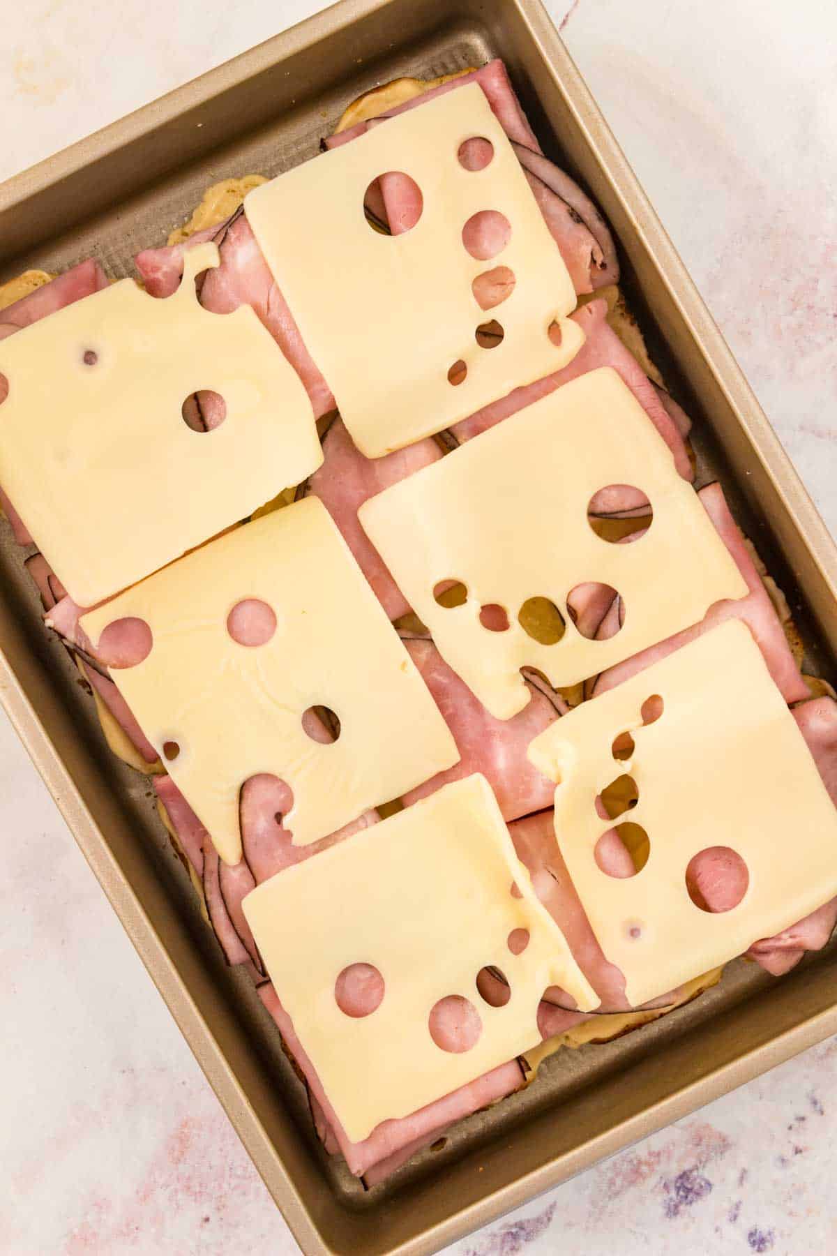 Slices of Swiss cheese added over ham sliders in a baking pan.