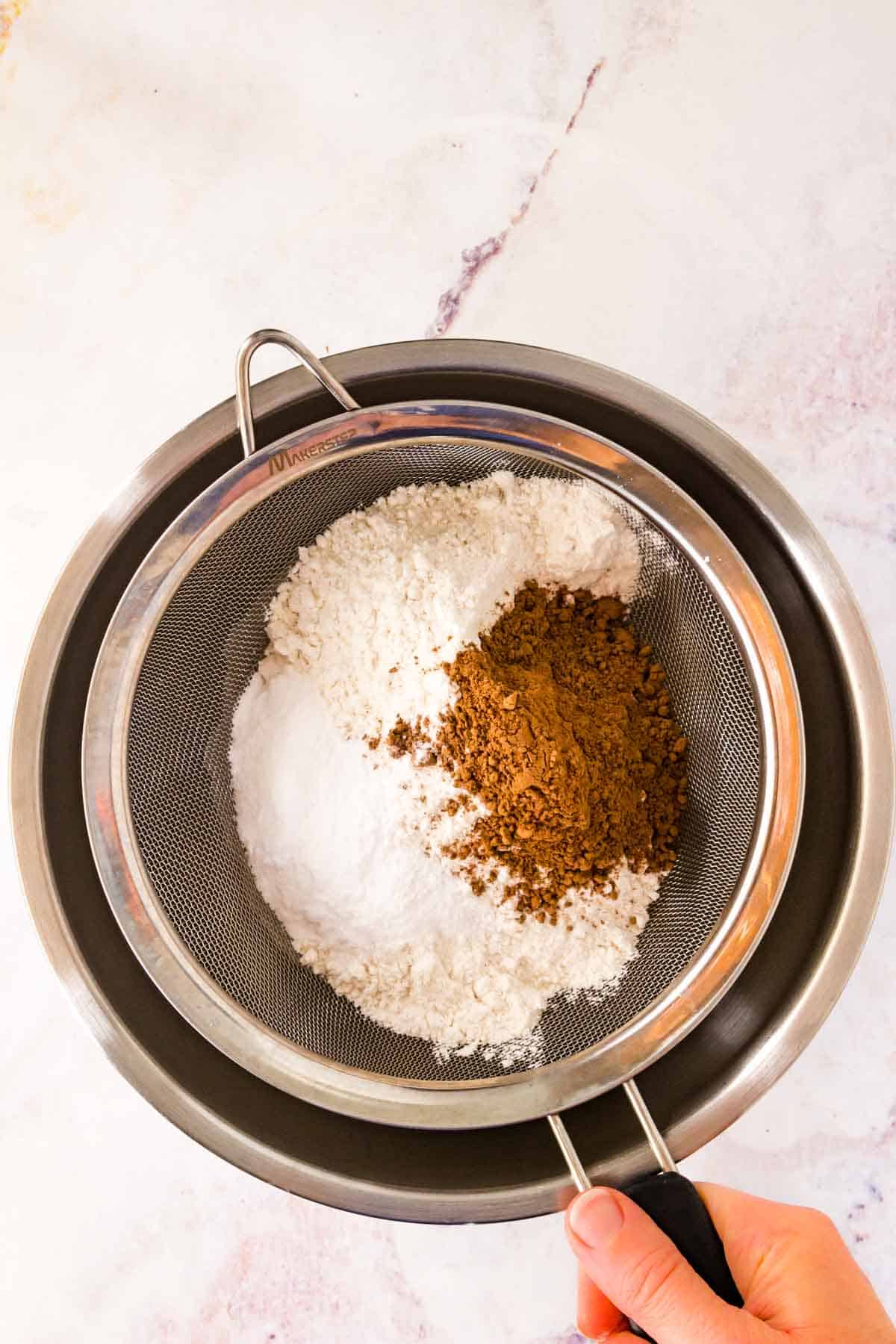 Dry ingredients sifting through a fine mesh sieve into a mixing bowl.