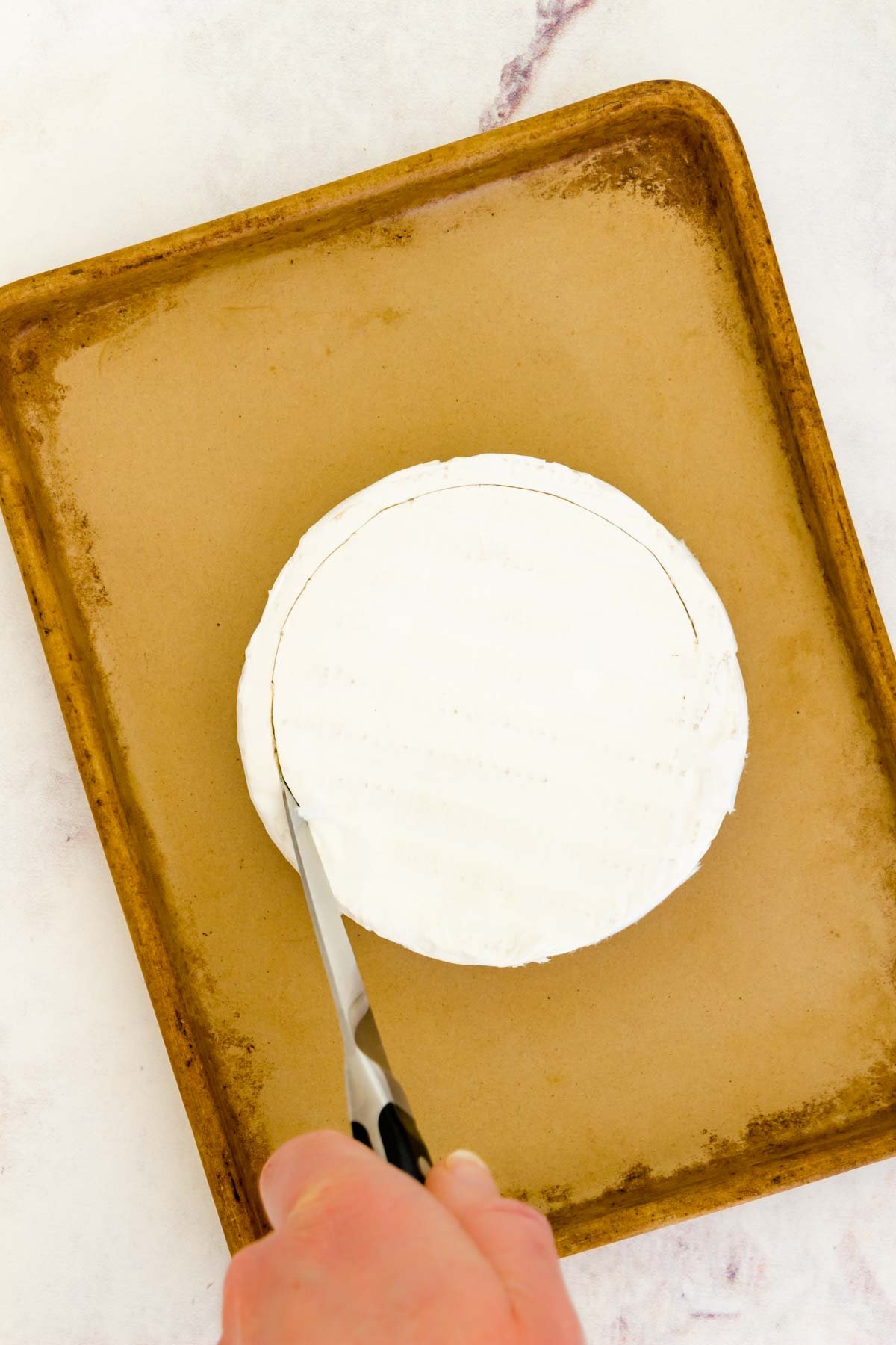 A knife scoring a circle around the edge of a wheel of Brie on a stoneware baking pan.