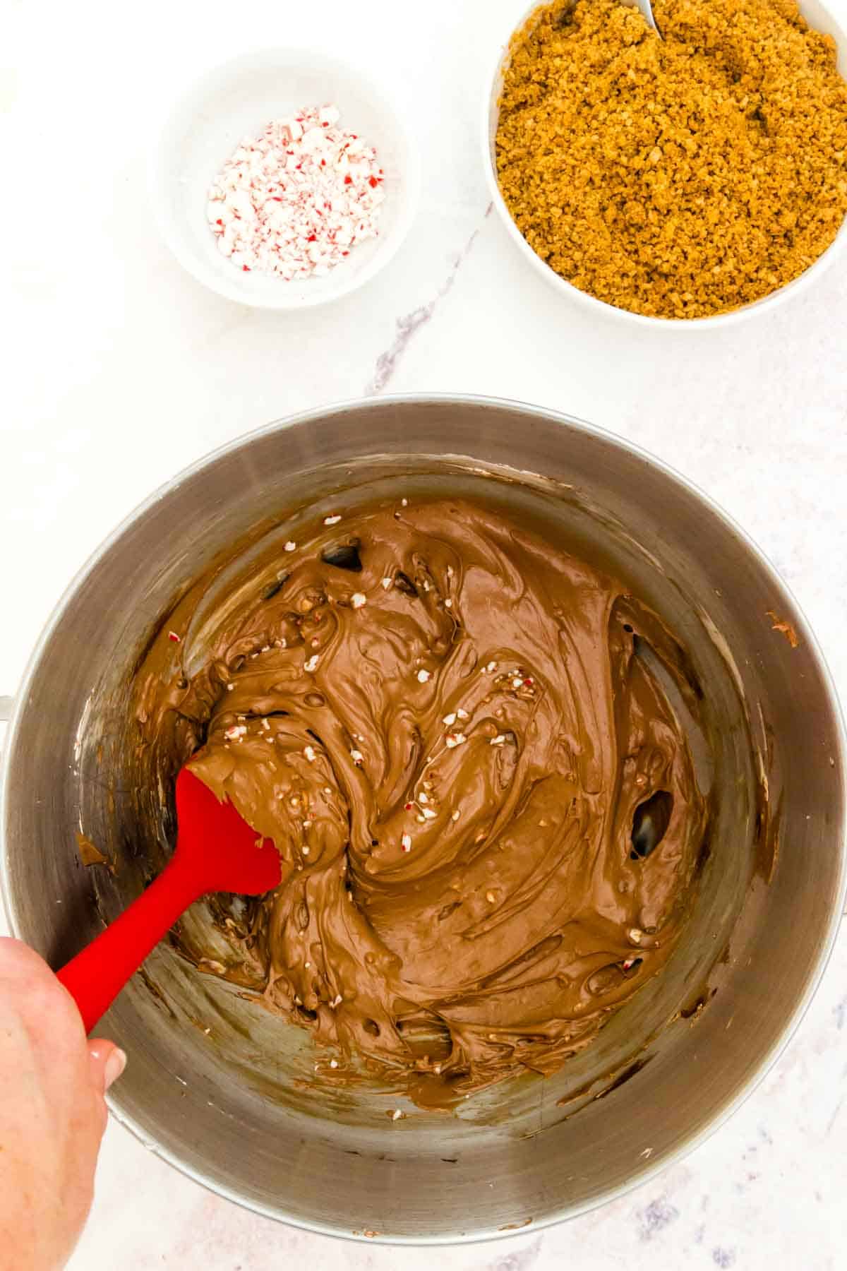 Stirring chopped candy canes into the chocolate cheesecake mixture with a red spatula in a large mixing bowl next to bowls of graham cracker crust mixture and chopped peppermint candies.