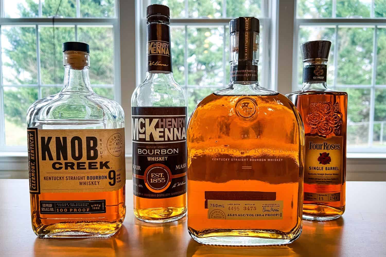 Bottles of Knob Creek, Henry McKenna, Woodford Reserve, and Four Roses Bourbon on a table in front of a window.