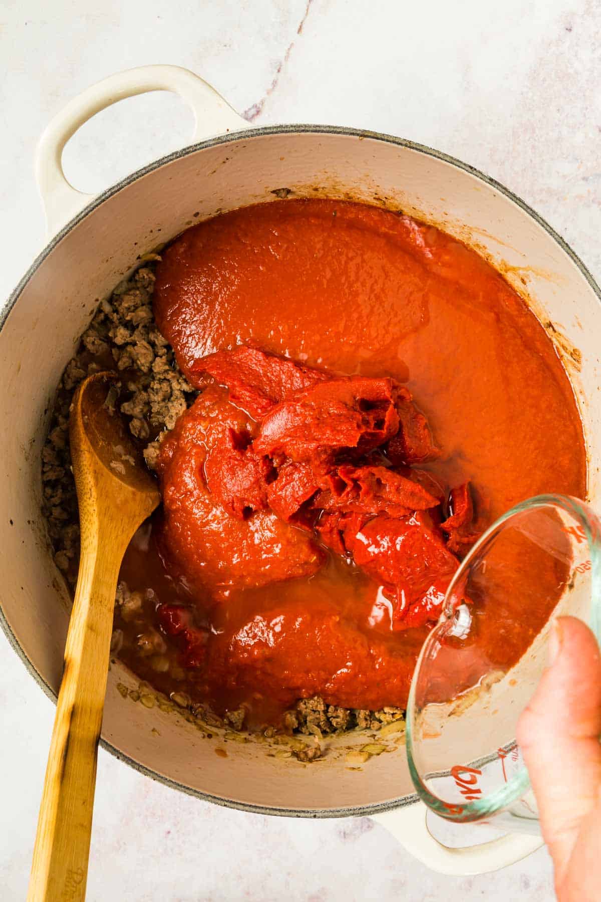 Water being poured into a pot with browned meat, tomato sauce, and tomato paste.