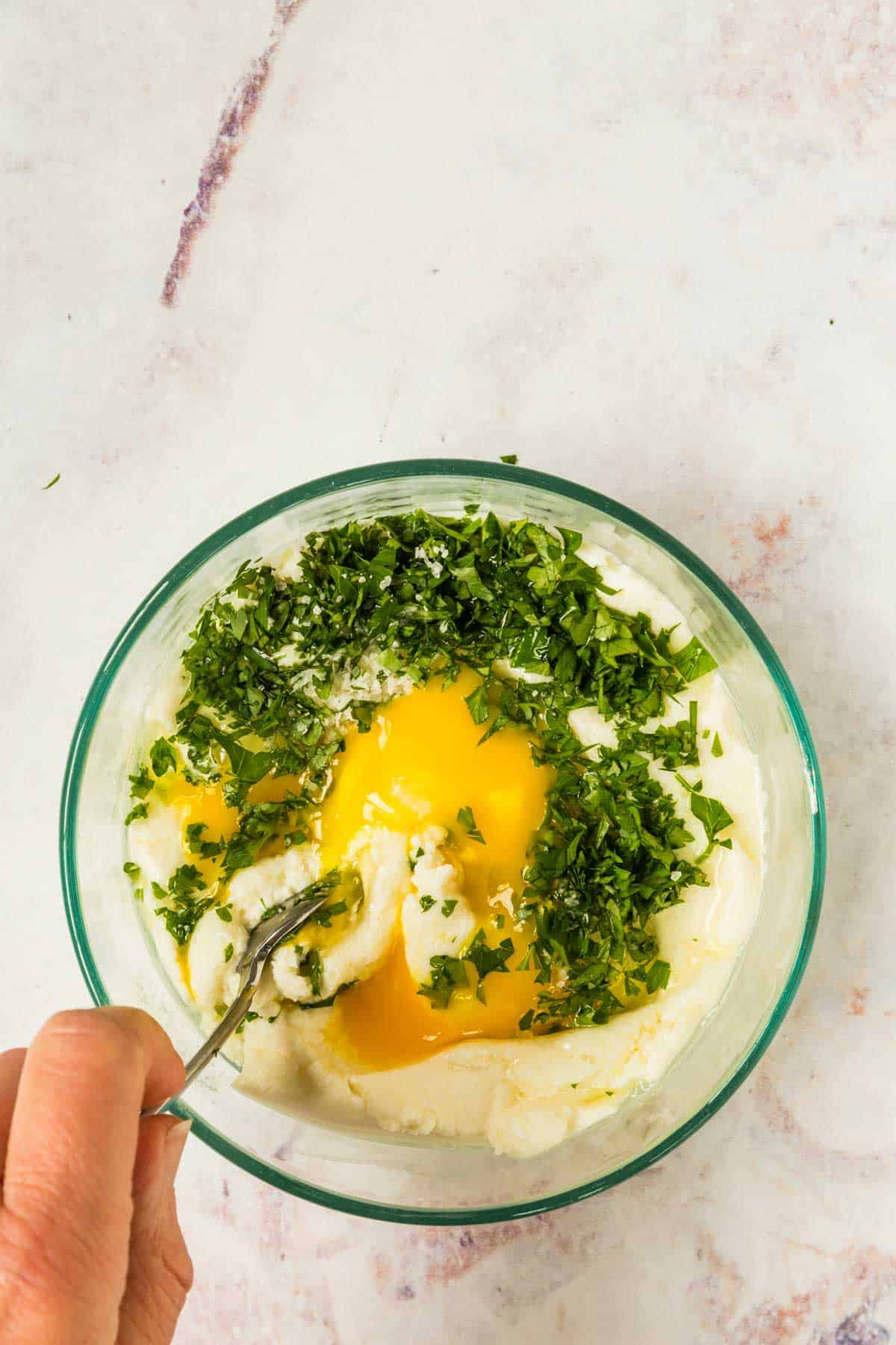 An egg and parsley being stirred into a bowl of ricotta cheese.