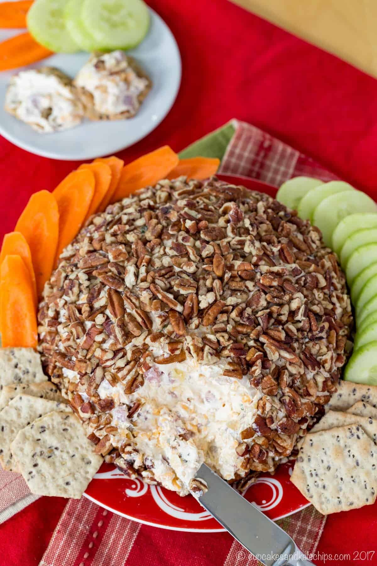 Looking down at cheeseball coated in pecans on a plate surrounded by crackers and raw vegetables with a small plate with two crackers topped with the ham and cheese spread next to it.