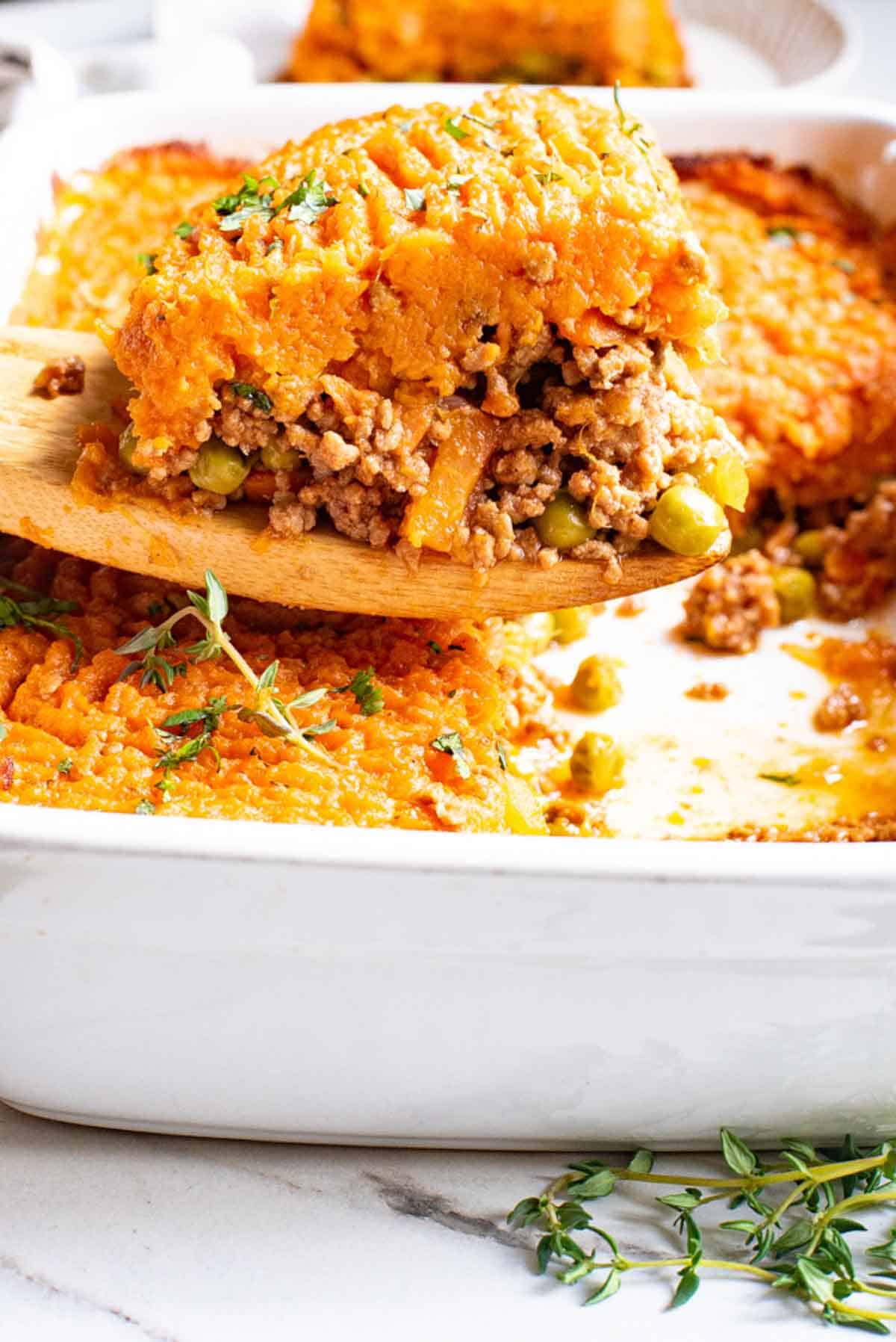 A serving of sweet potato shepherd's pie is lifted out of a casserole dish with a wooden spoon.