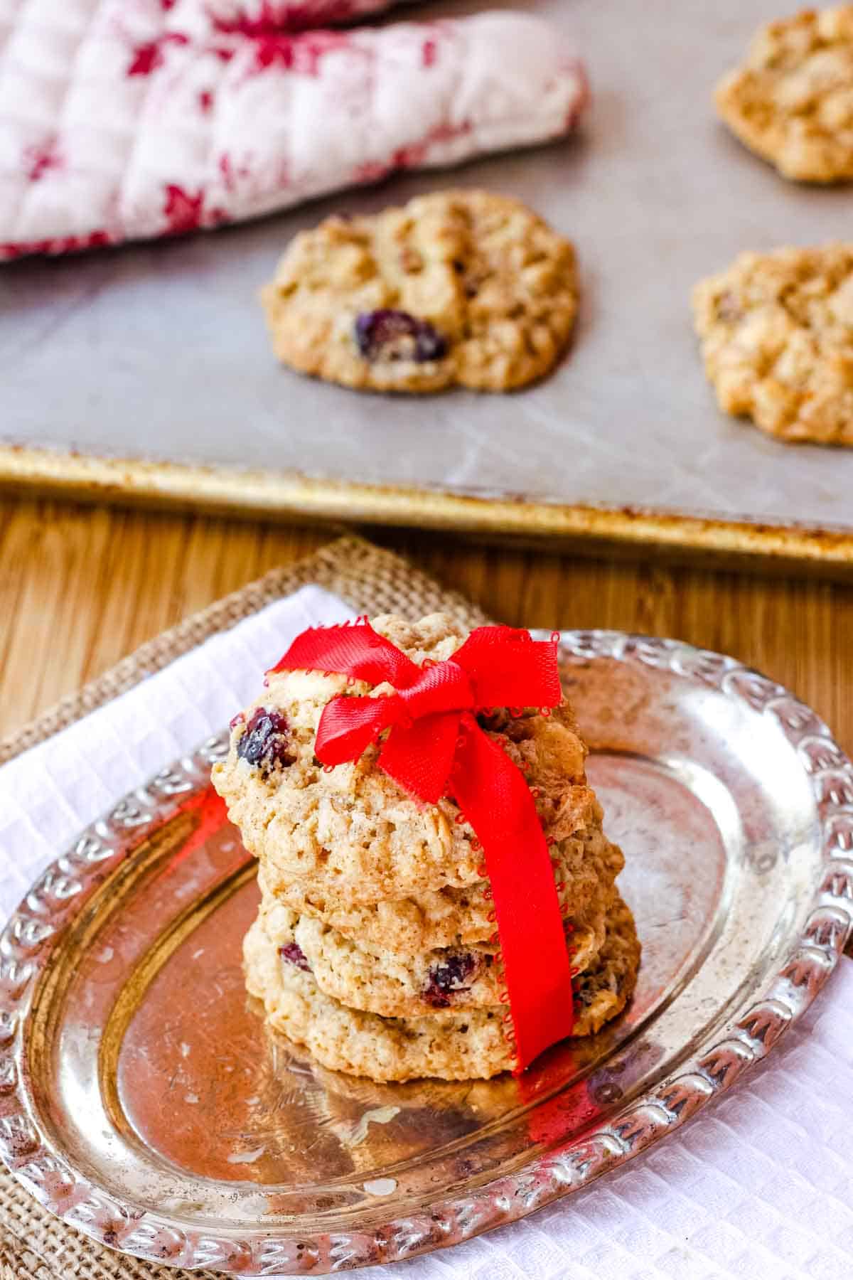 Four White Chocolate Cranberry Oatmeal Cookies tied with a red ribbon ona silver oval dish and more on a cookie sheet behind them with an oven mitt.