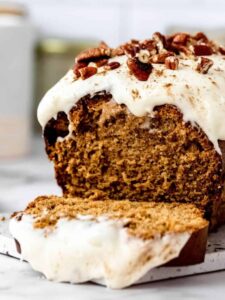 A gingerbread loaf with cream cheese frosting on a marble cutting board with the first slice cut off and laying in front of it.