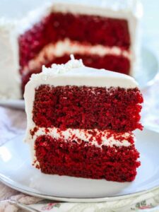 A slice of gluten-free red velvet cake with cream cheese frosting on a white plate with the rest of the cake in the background.