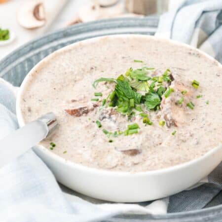 A bowl of cream of mushroom soup with a spoon in it garnished with fresh parsley, on a cloth napkin in a metal tray.