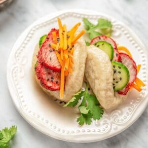 Two gluten free bao buns filled with char siu pork, shredded carrots, and cucumber slices on a plate garnished with cilantro.