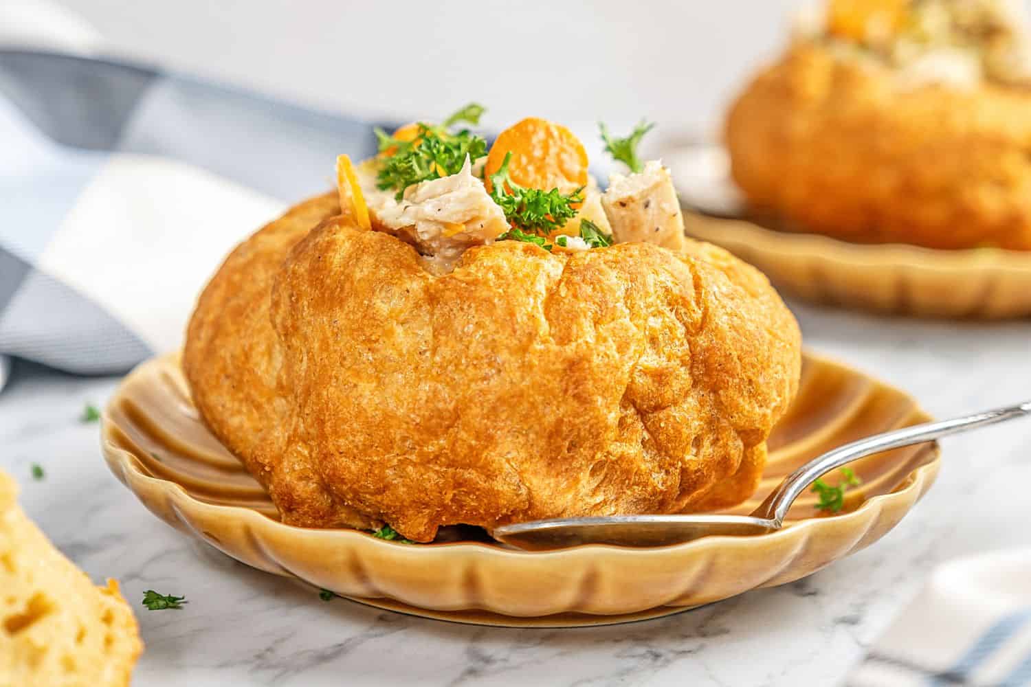 A gluten-free bread bowl on a tan scallop-edged plate with a spoon filled with chicken and vegetables.