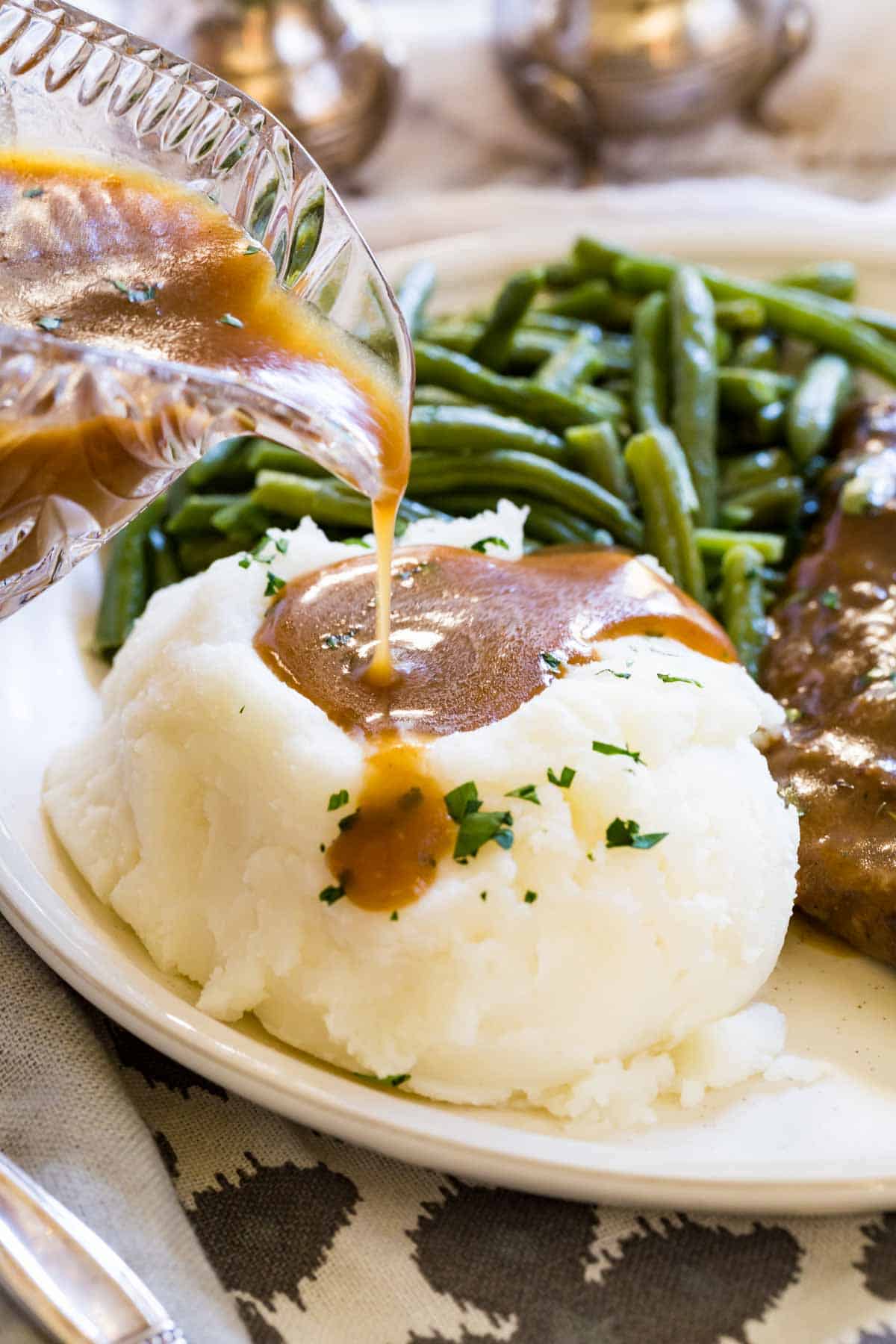 Gluten-free brown gravy is poured over a pile of mashed potatoes on a plate next to green beans.