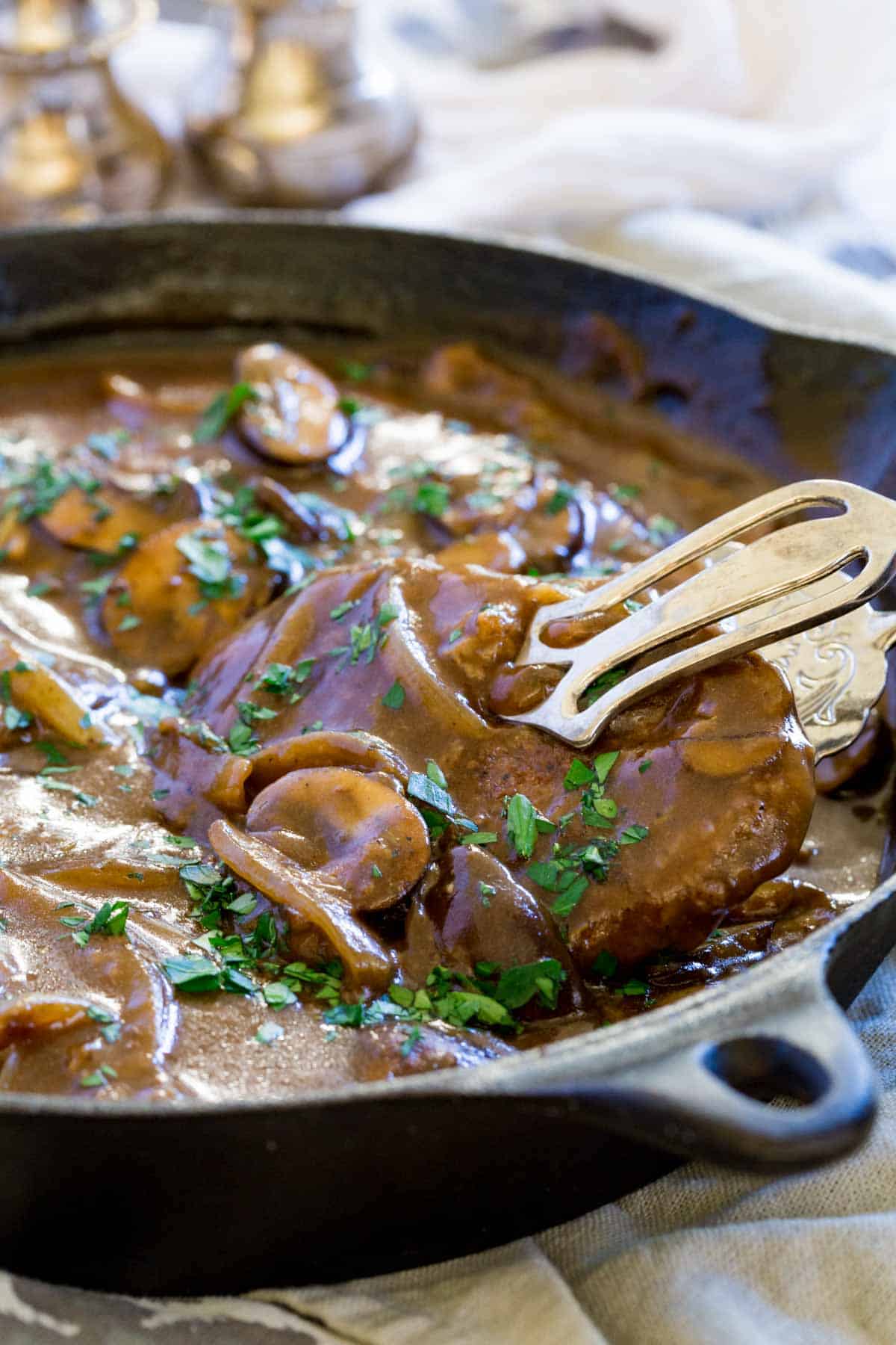 Tongs lifting a cube steak from a skillet with gravy and mushrooms.