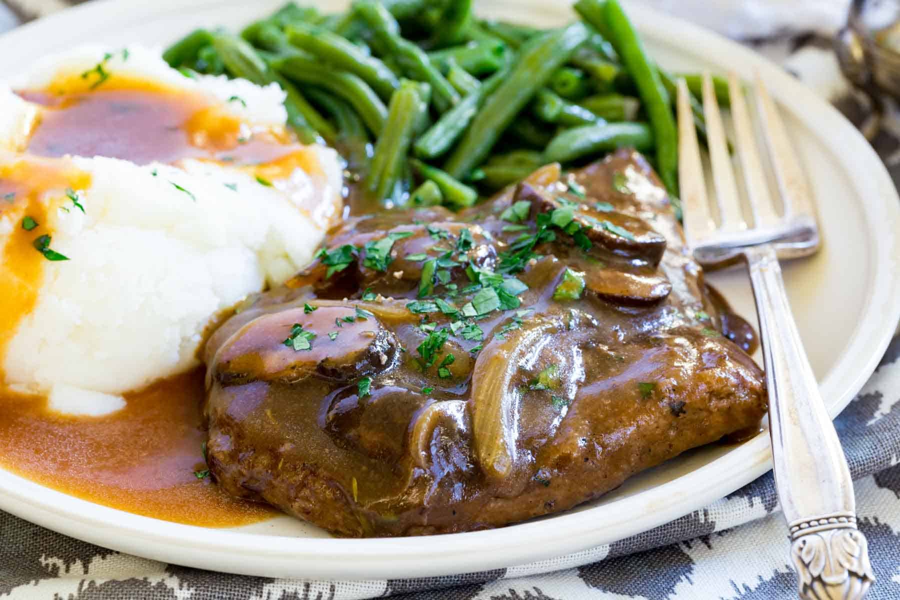 Gluten-free cube steak with gravy topped with mushrooms and onions on a plate next to mashed potatoes and green beans, with a fork.