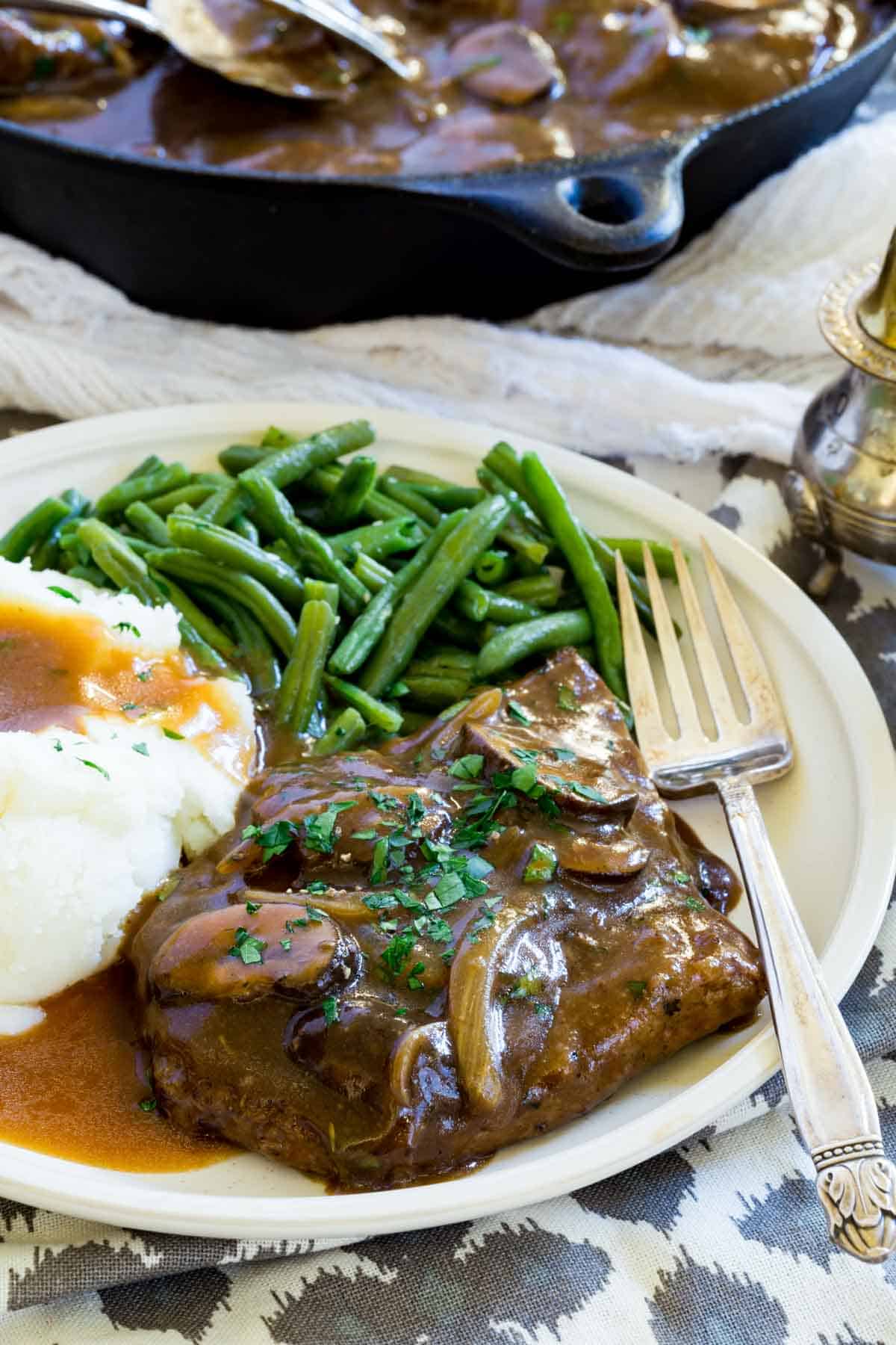 Gluten-free cube steak with gravy topped with mushrooms and onions on a plate next to mashed potatoes and green beans, with a fork.