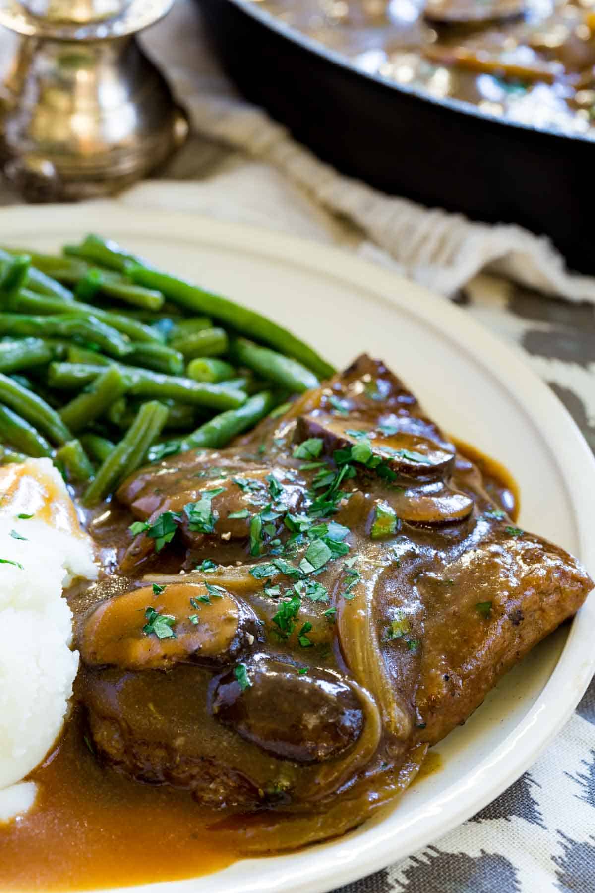 Gluten-free cube steak with gravy topped with mushrooms and onions on a plate next to mashed potatoes and green beans, with a fork.