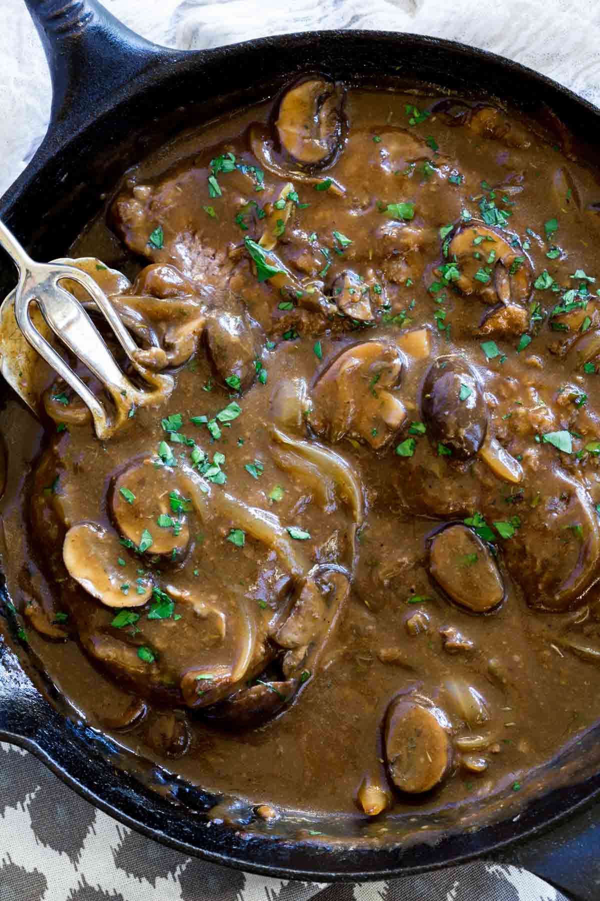 Overhead view of tongs lifting a cube steak from a skillet with gravy and mushrooms.