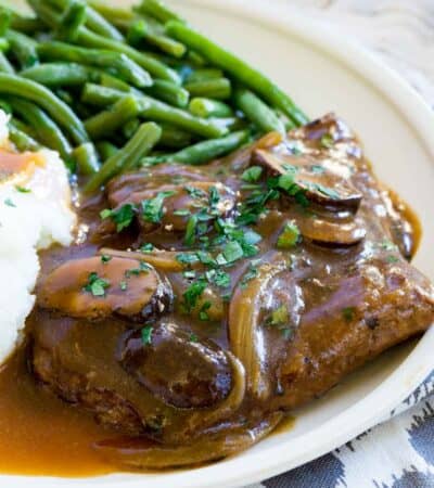 Gluten-free cube steak with gravy topped with mushrooms and onions on a plate next to mashed potatoes and green beans.