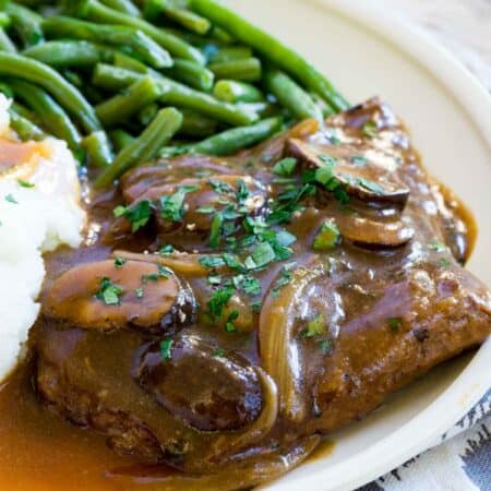 Gluten-free cube steak with gravy topped with mushrooms and onions on a plate next to mashed potatoes and green beans.