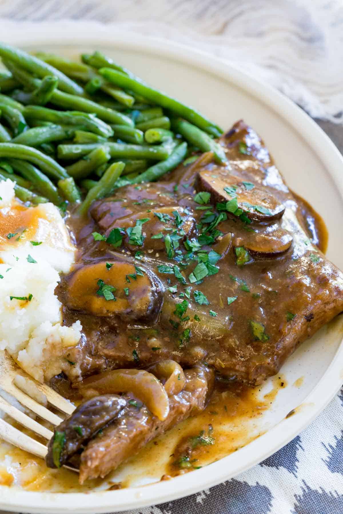 Gluten-free cube steak with gravy topped with mushrooms and onions on a plate next to mashed potatoes and green beans, with a fork.