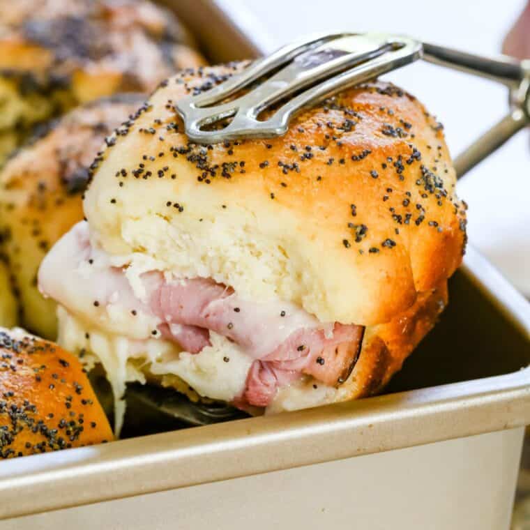 A ham and cheese slider being picked up out of a baking pan with serving tongs.