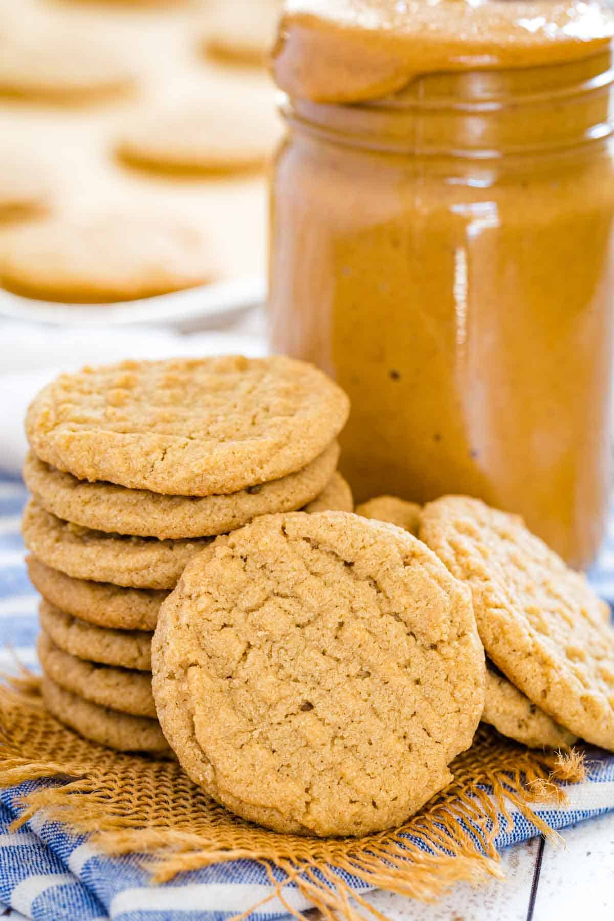 A gluten-free peanut butter cookie leaning against a stack of peanut butter cookies, with a jar of peanut butter in the background.