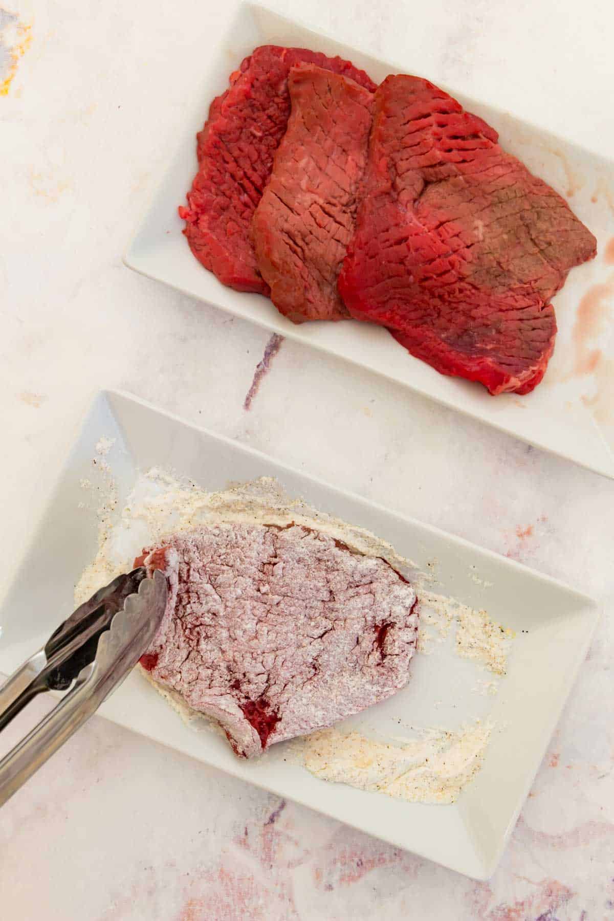 Tongs are used to dredge a piece of cube steak through a seasoned flour mixture, next to a plate of cube steaks.