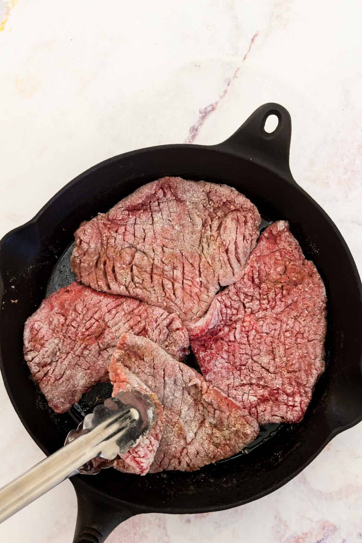 Tongs are used to flip one cube steak out of four that are searing in a cast iron skillet.
