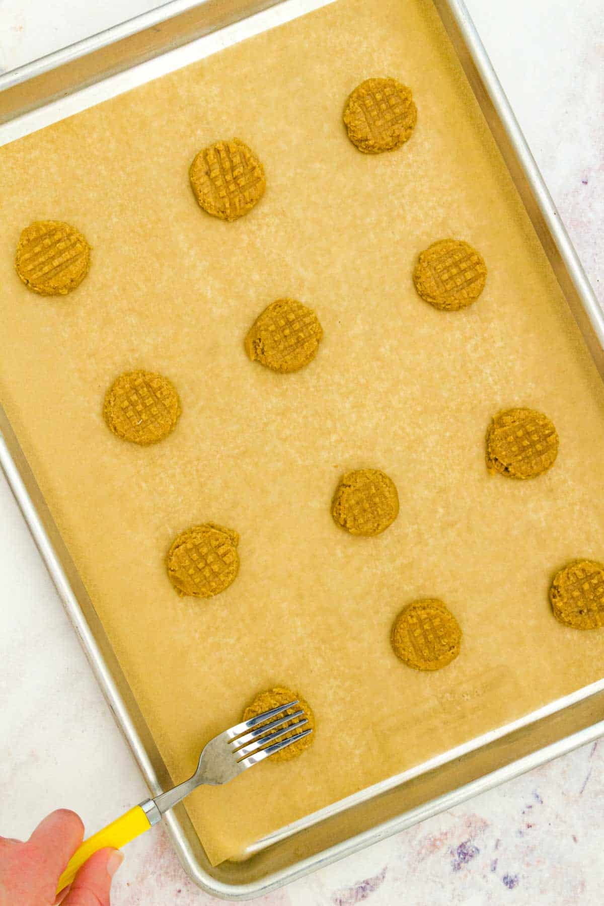 A hand using a fork to press indents into the tops of gluten-free peanut butter cookie dough on a baking sheet.