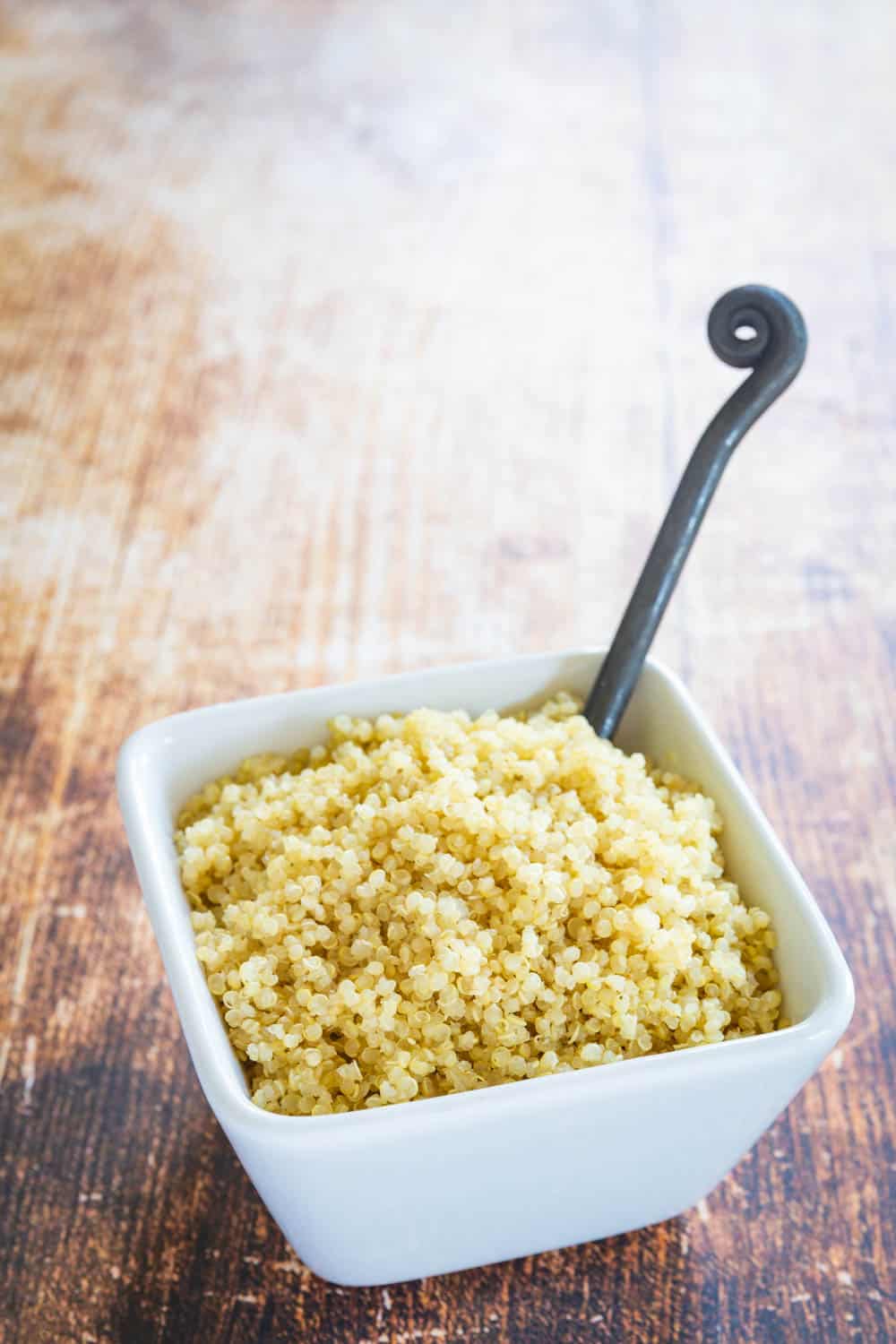 A white ceramic bowl of cooked quinoa with a serving spoon in it.