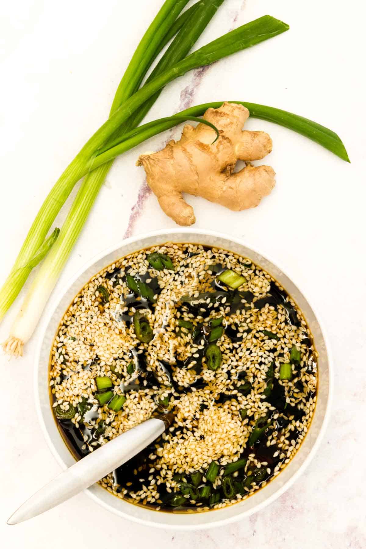 A bowl of the sesame glaze sauce on a tabletop next to a couple of whole scallions and a knob of fresh ginger.
