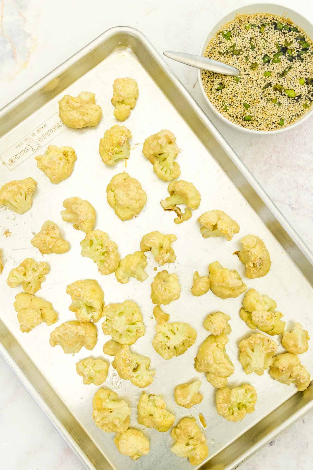 Partially baked cauliflower bites on a baking sheet with the bowl of sesame glaze next to it.