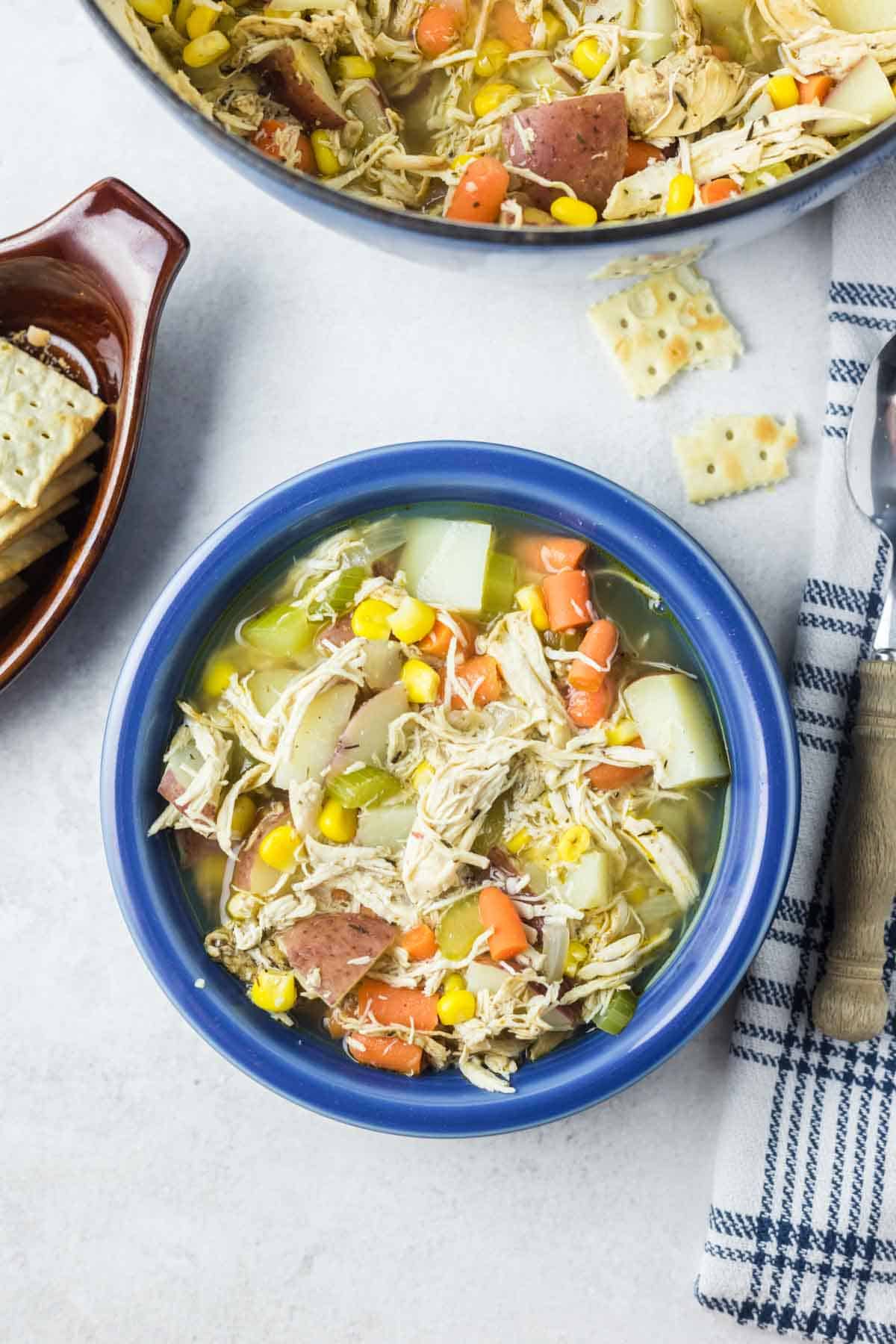 A serving of rotisserie chicken soup in a blue bowl, next to a large pot of soup.