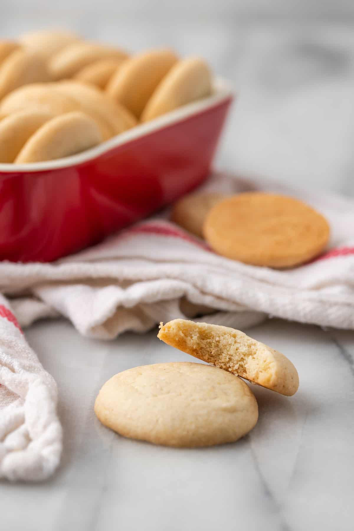 Two gluten-free vanilla wafers stacked on top of one another, with a bite missing from the top cookie, and more cookies in a baking dish in the background.